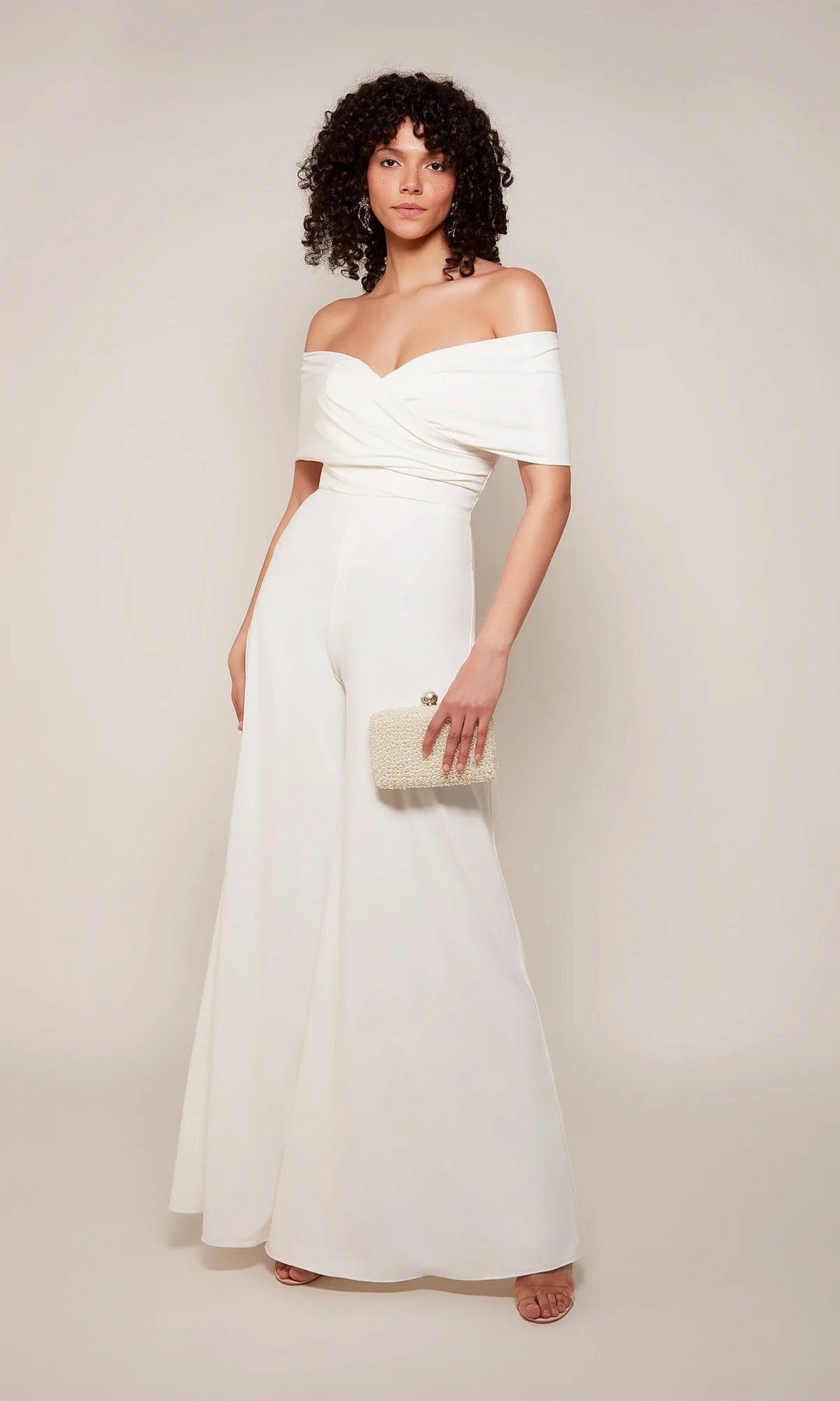 A woman stands against a plain background wearing the Alyce 70051 long white jumpsuit in Diamond White and holding a small beaded clutch.